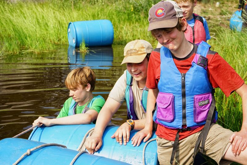 The image shows four young people on a raft made of blue barrels in a body of water. Three of them are wearing life vests. The person in the front is holding a rope. There is a blue barrel on the shore in the background, along with green grass and trees. The raft appears to be homemade.